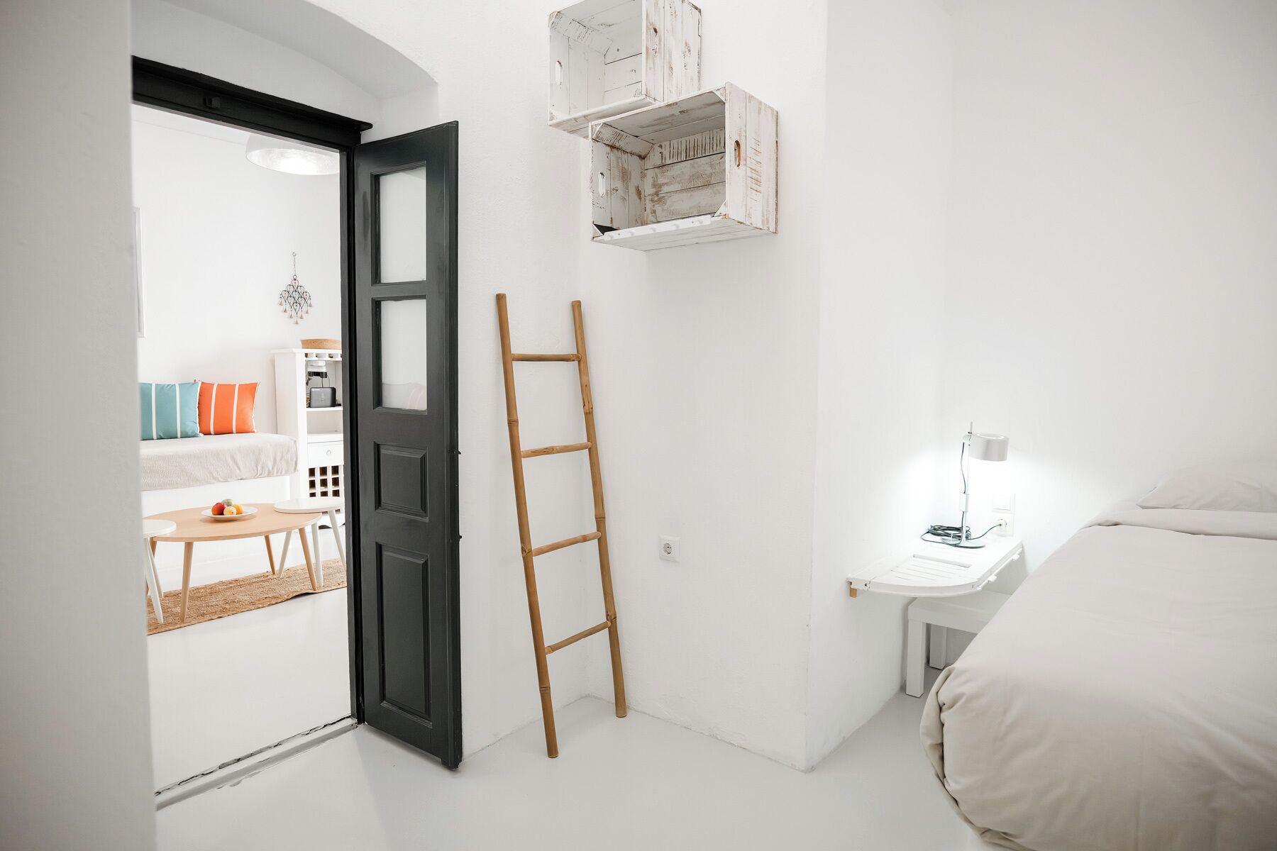 Un dormitorio con cama, una mesita de noche con lámpara, una escalera apoyada en la pared y una puerta que da a otra habitación.