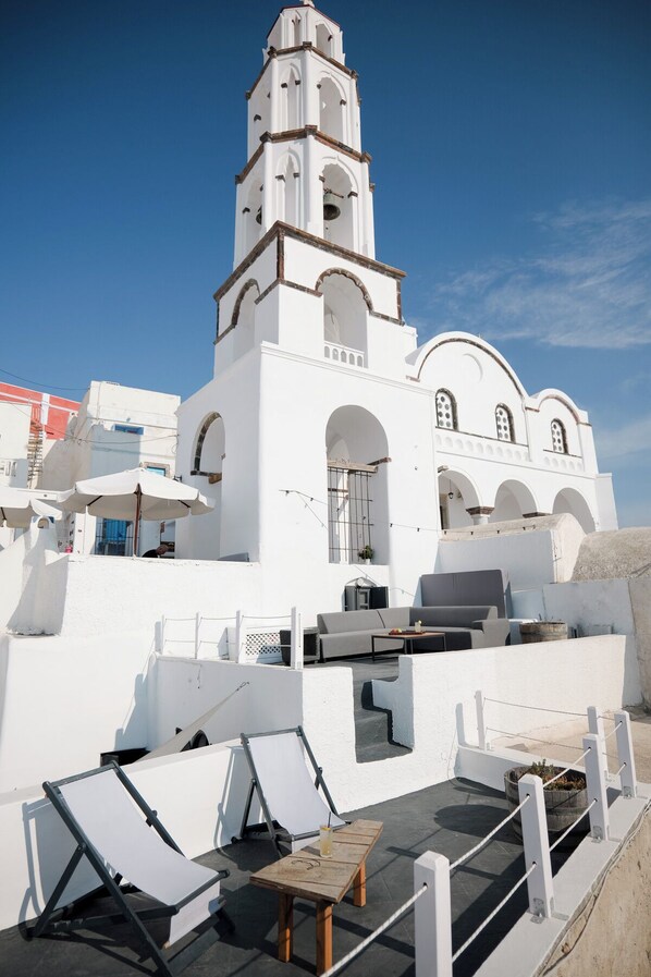 A white building with a bell tower and arched windows, featuring outdoor seating and umbrellas.