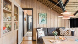 Una habitación acogedora con paredes de madera, un sofá con cojines de lunares, una mesa de comedor con un jarrón de flores y un aparador con puertas de cristal.