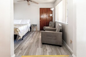 A bedroom with a bed, a chair, a small table, a wooden door, and a window.