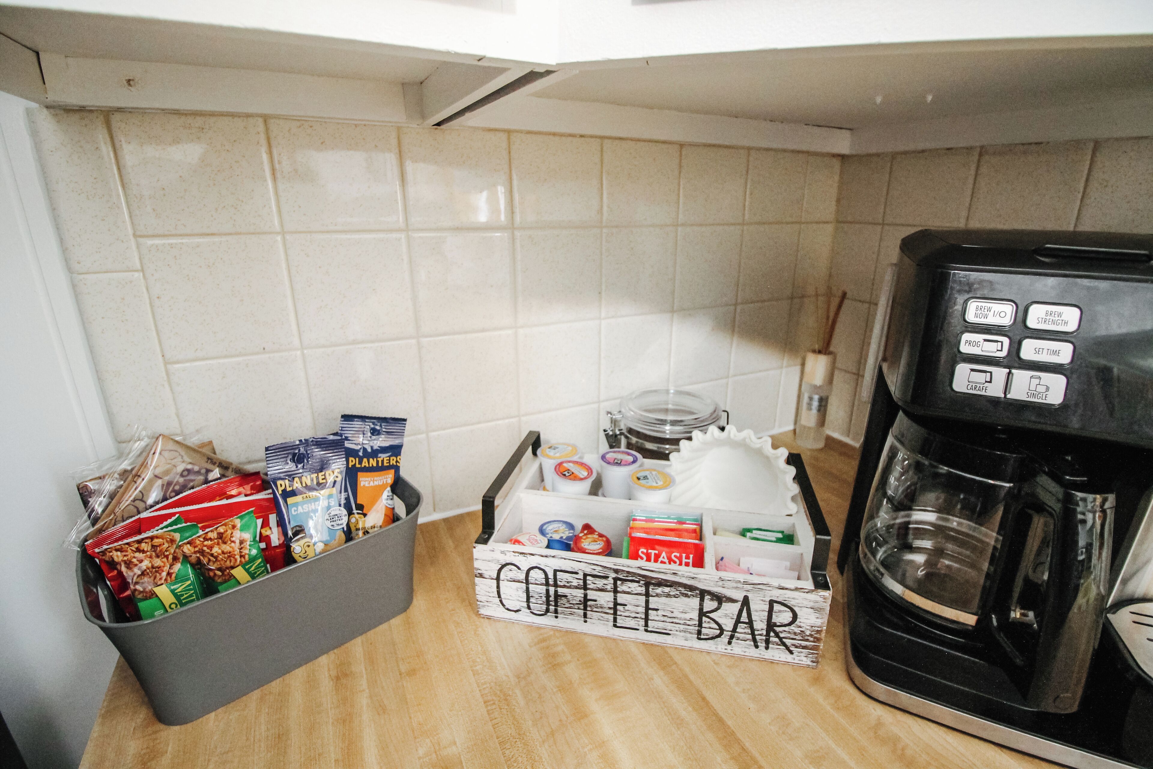 Et tekøkken med kaffemaskine, en opbevaringskasse med kaffekapsler og en mærket æske til kaffebarsartikler.