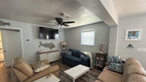 A living room with a ceiling fan, a TV, a sofa, a coffee table, and a window with blinds. - Sargent Barefoot Bungalow (Bay City)