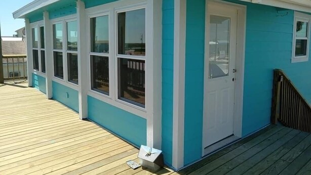 A small, single-story building with a light blue exterior and white trim. It features a front door, multiple windows, and a wooden deck. - Sargent Barefoot Bungalow (Bay City)
