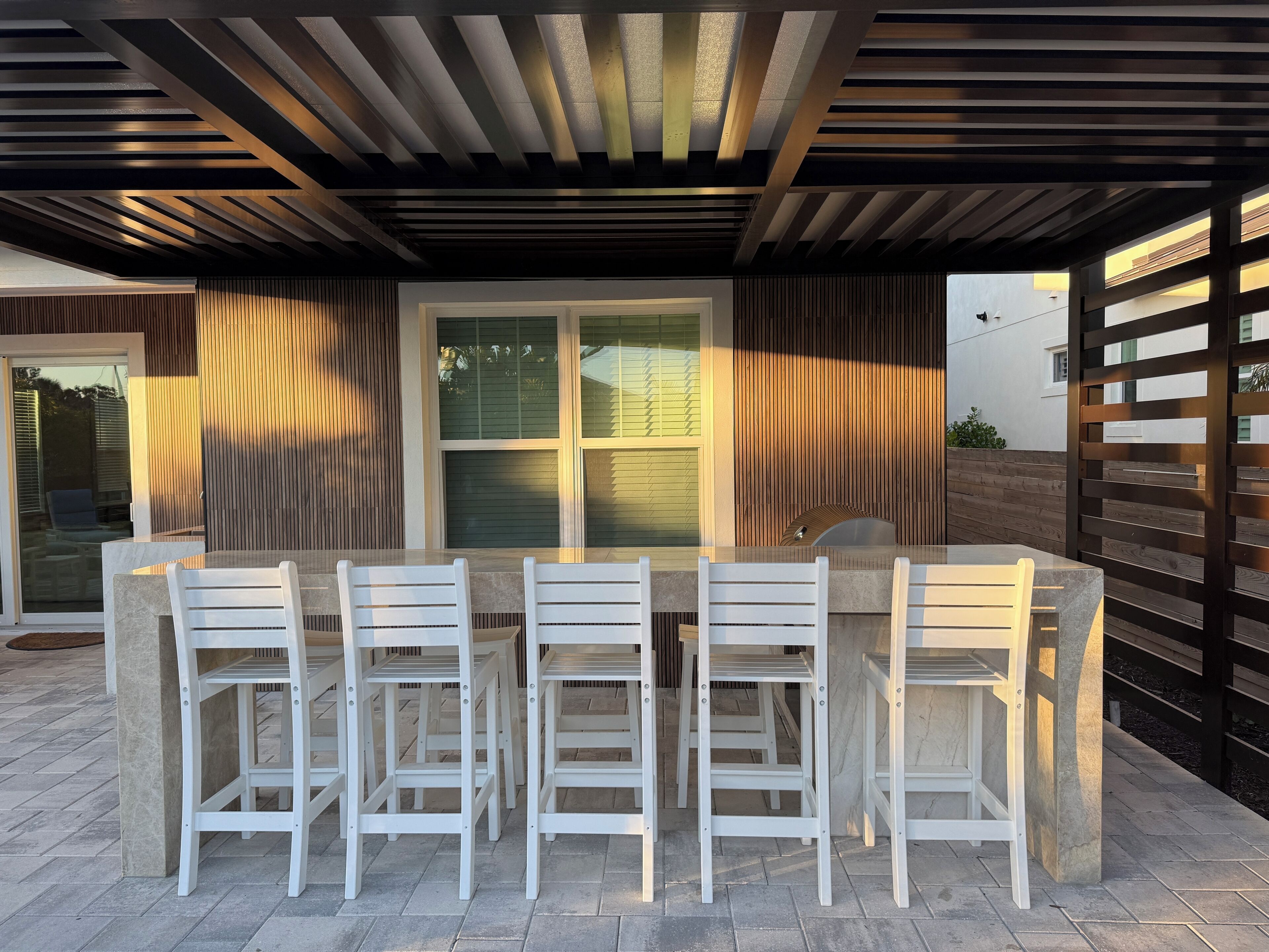 Une terrasse extérieure avec une table en pierre et quatre chaises blanches, entourée de lattes de bois et d’une porte vitrée.