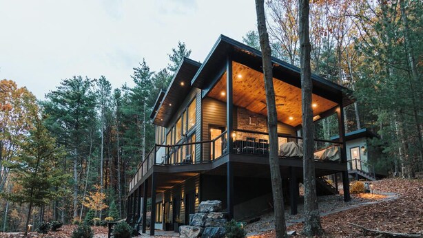 A modern log cabin with a large deck and multiple windows, surrounded by a forested landscape. - TreeTops, modern luxury overlooking Keuka Lake - Luxury Treehouse included (Penn Yan)