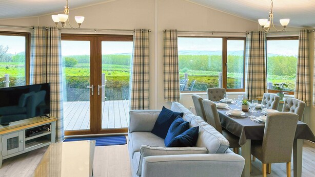Ein geräumiges Wohnzimmer mit einer Couch, Fernseher und einem Essbereich mit Stühlen und Tisch. Große Fenster bieten Blick auf eine grüne Landschaft.