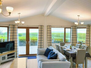 Ein geräumiges Wohnzimmer mit einer Couch, Fernseher und einem Essbereich mit Stühlen und Tisch. Große Fenster bieten Blick auf eine grüne Landschaft.