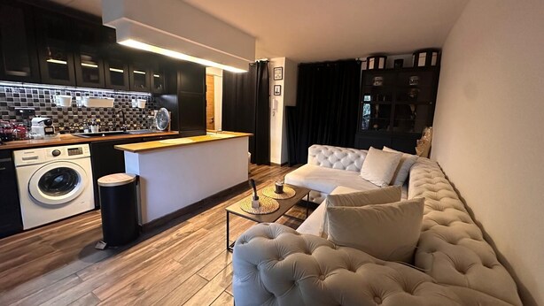 Eine moderne Küche mit integriertem Waschtrockner, einer Kaffeemaschine und einer Holz Arbeitsplatte. Im Wohnbereich befindet sich ein weißes Sofa mit einer gesteppten Rückenlehne sowie ein kleiner Couchtisch.