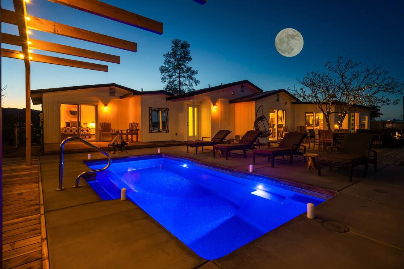 Ein Poolbereich mit Liegestühlen und einer Holzterrasse, ein Haus im Hintergrund mit einem Mond, und ein Baum.