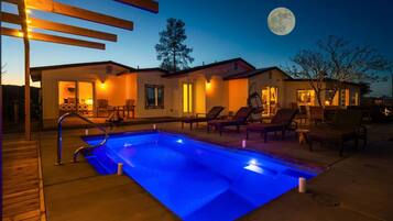 Ein Poolbereich mit Liegestühlen und einer Holzterrasse, ein Haus im Hintergrund mit einem Mond, und ein Baum.