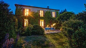 A stone building with ivy covering the walls, a well-lit outdoor area with a table and chairs, and a clear sky. - Beautiful Stone Farmhouse, Private Pool, Idyllic Setting Amongst Vineyards near Sainte-Foy-la-Grande (Sainte Foy la Grande)