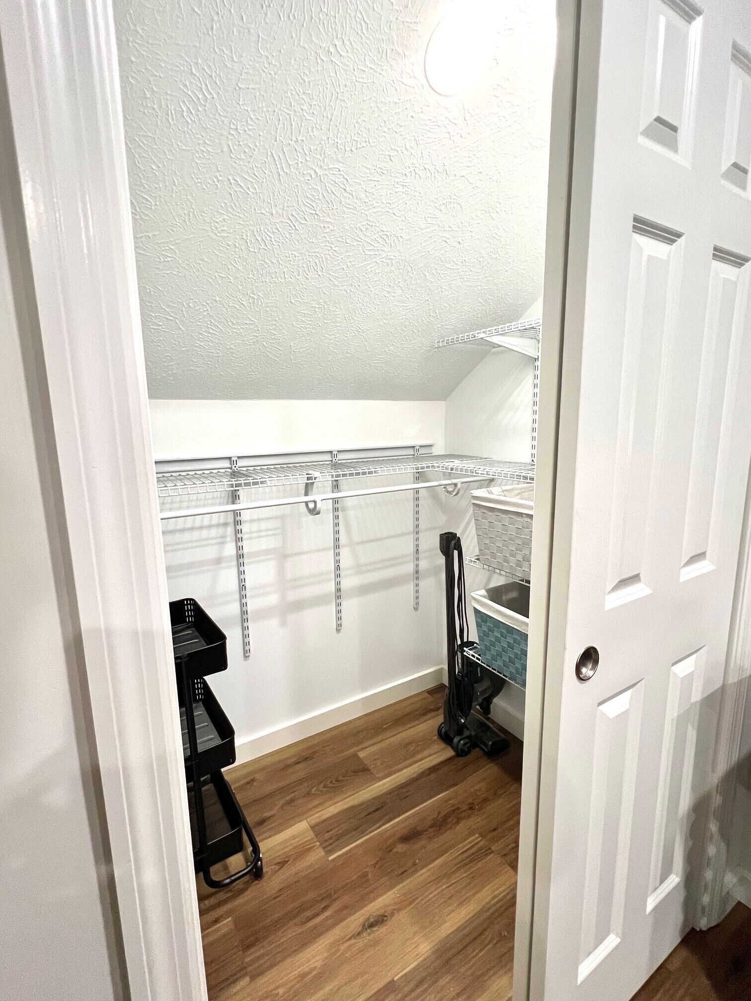 A small, narrow room with a white door, a white ceiling, and a wooden floor. There is a black stool and a black chair in the corner.