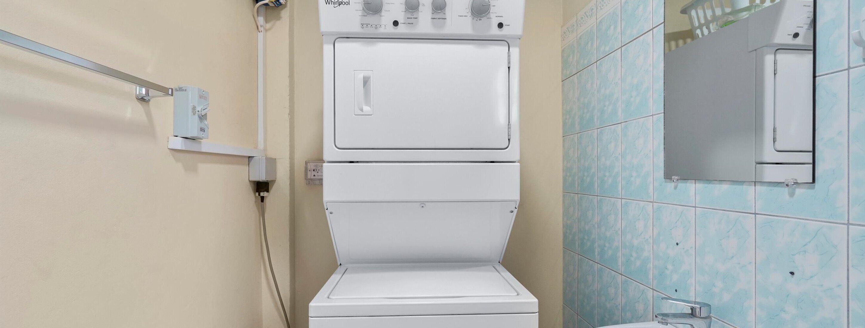 A compact, two-tiered washer-dryer unit in a bathroom with light blue tiles and a beige wall.