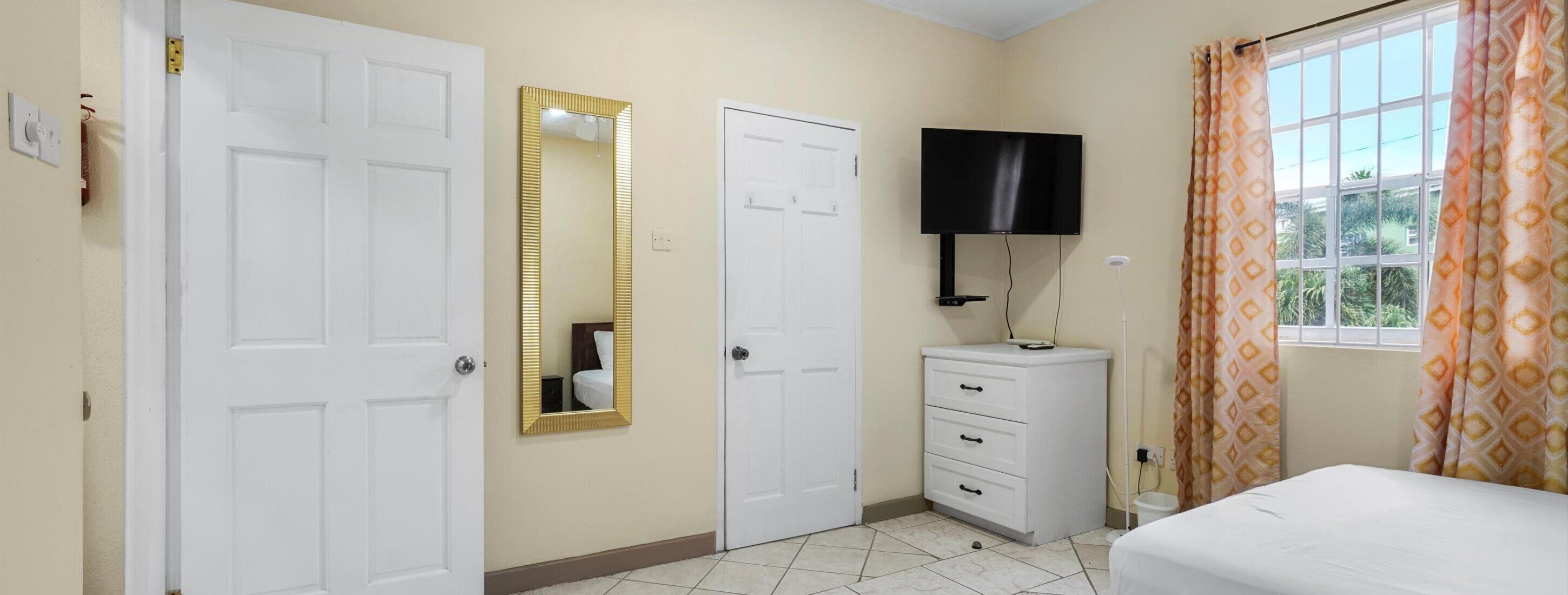Un dormitorio con dos puertas blancas, un tocador blanco y un televisor de pantalla plana montado encima, además de una cama con cubrecama blanco.