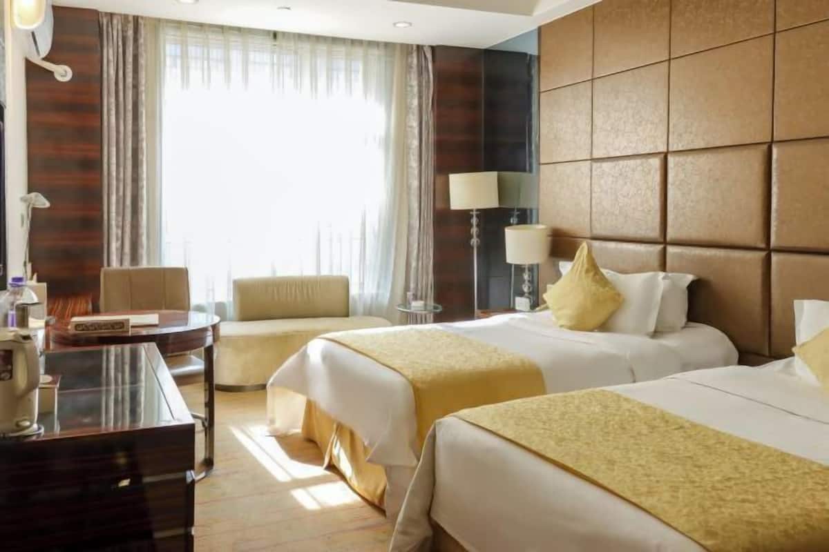 Quarto de hotel com duas camas, uma escrivaninha, uma cadeira e uma janela grande com cortinas.