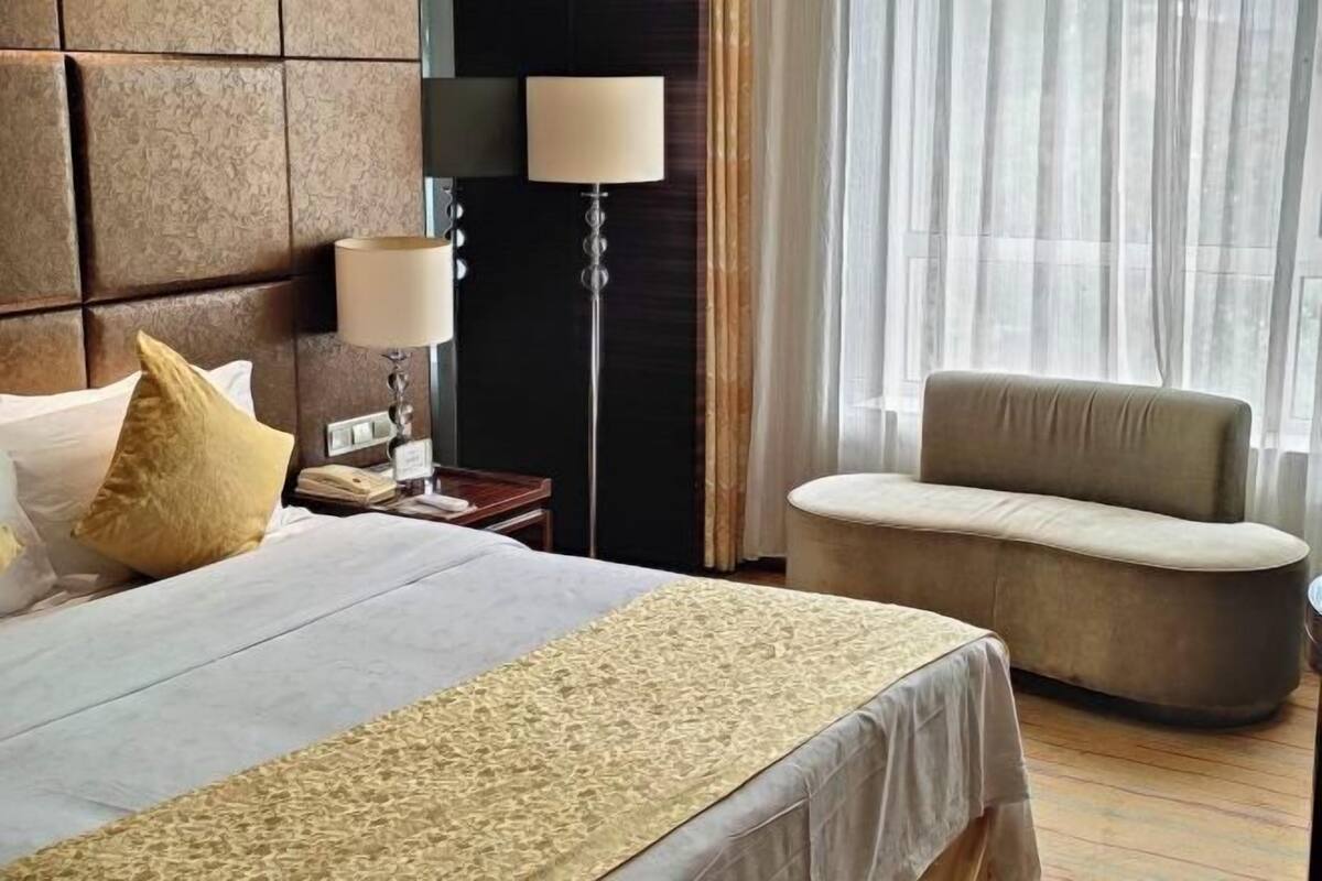 Une chambre d’hôtel avec un grand lit, une table de chevet, une lampe, une chaise et une vue sur l’extérieur à travers des rideaux légers.