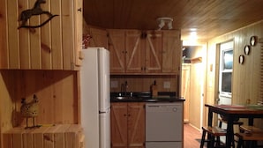 Ett kök med träinredning, kylskåp, diskmaskin och träskåp. Där finns ett litet bord med stolar och en dekorativ kycklingfigur.