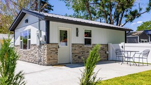 A small, single-story building with a stone foundation and white siding, featuring a door, windows, and a small patio area with outdoor seating. - Beautiful NEW 1-bed, 1-bath right in downtown Cody! Sleeps up to 4.  (Cody)