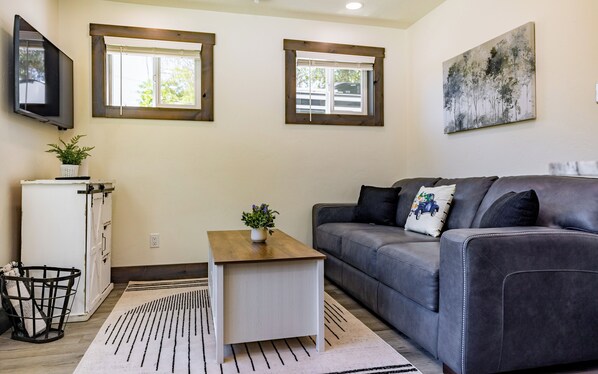 Una sala de estar con un sofá gris, una mesa de centro de madera, un armario blanco y dos ventanas con persianas.