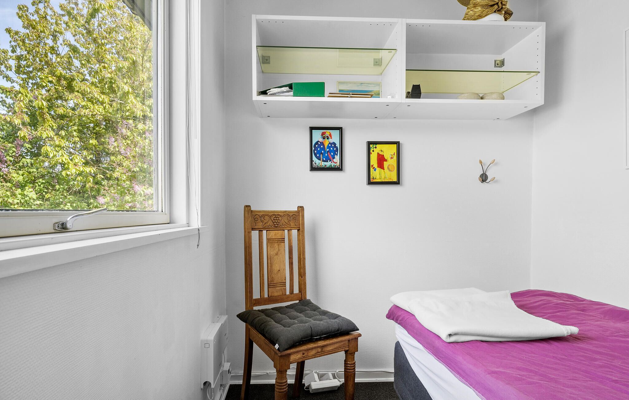 Ein Schlafzimmer mit einem hölzernen Stuhl, einem Bett mit lila Bettdecke, einem weißen Regal über dem Bett und zwei gerahmten Bildern an der Wand.