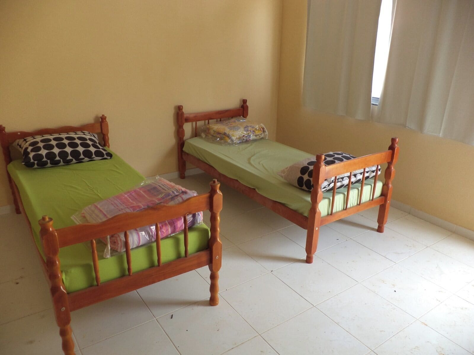 Dos camas de madera con colchones verdes y almohadas de lunares en una habitación con paredes amarillas y una ventana con persianas.