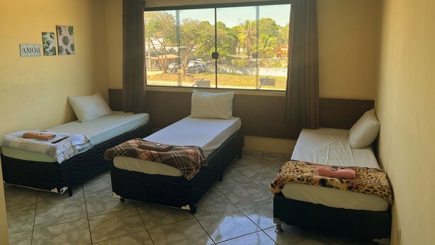 A room with three beds arranged in a row, a large window with a view of the outside, and a ceiling fan. - Hotel Ita Avenida (Itaborai)