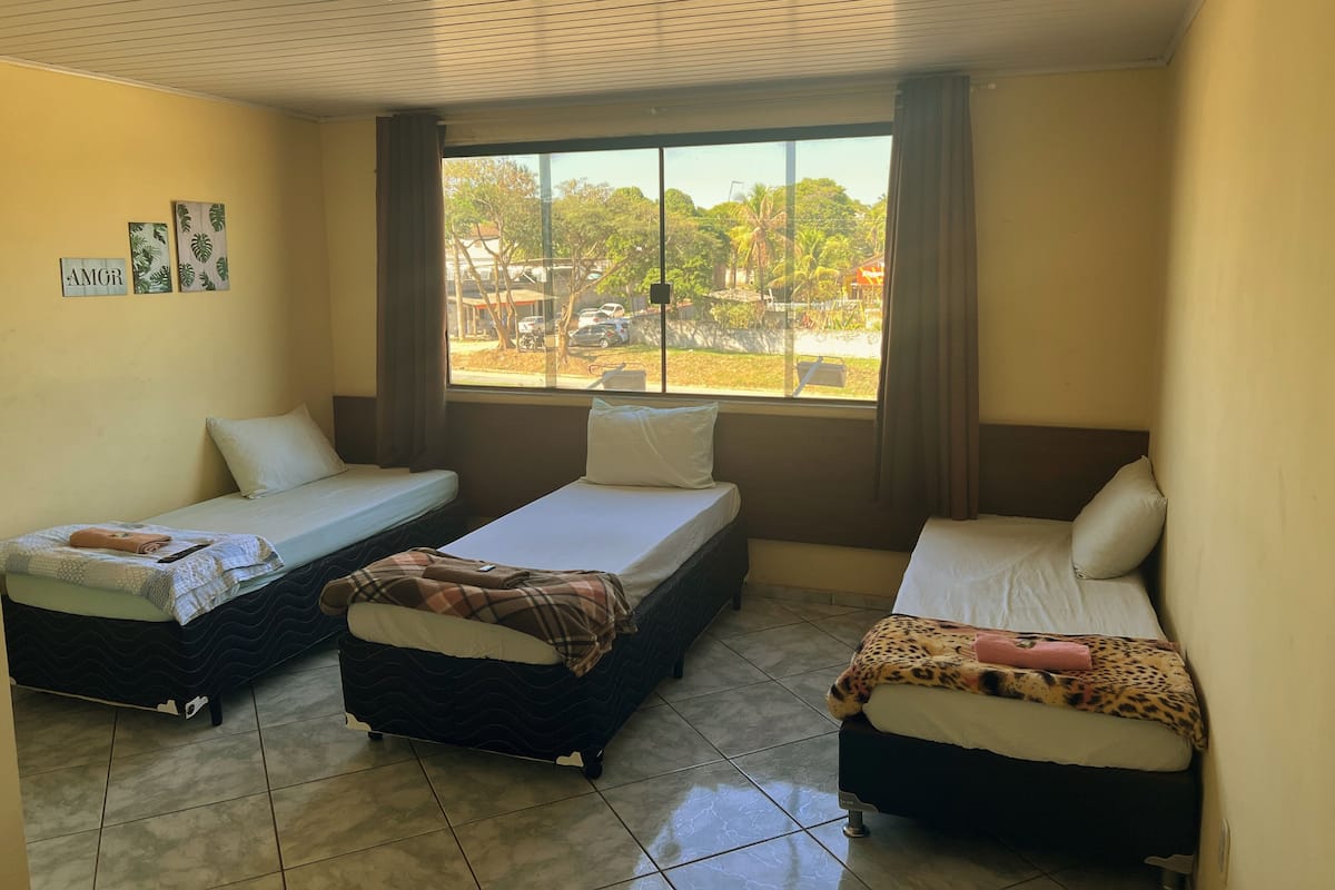 A room with three beds arranged in a row, a large window with a view of the outside, and a ceiling fan.