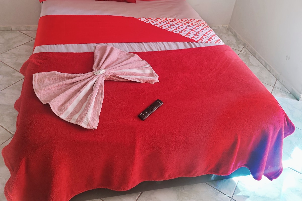 A bed with a red and white bedspread, a pink towel, and a remote control on the floor.
