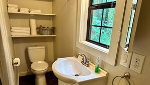 A bathroom with a toilet, sink, shelves with towels, and a window. - Meadows - A tiny touch of heaven at Stag Leap Cabins! (Nacogdoches)