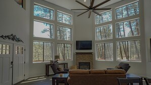 A spacious living room with a fireplace, large windows, and a ceiling fan. - OASIS IN THE FOREST ON 1.25 ACRE BACKING TO NATIONAL FOREST WITH AC-PETS WELCOME (Show Low)