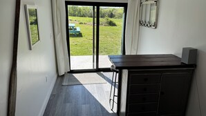 Een kamer met een donker houten bureau, een stoel, een klein kastje, een fotolijst en uitzicht op een groen buitengebied door een glazen schuifdeur.