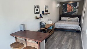 Een kleine kamer met een bed, een tafeltje met krukjes, een wandplank en een groot raam.