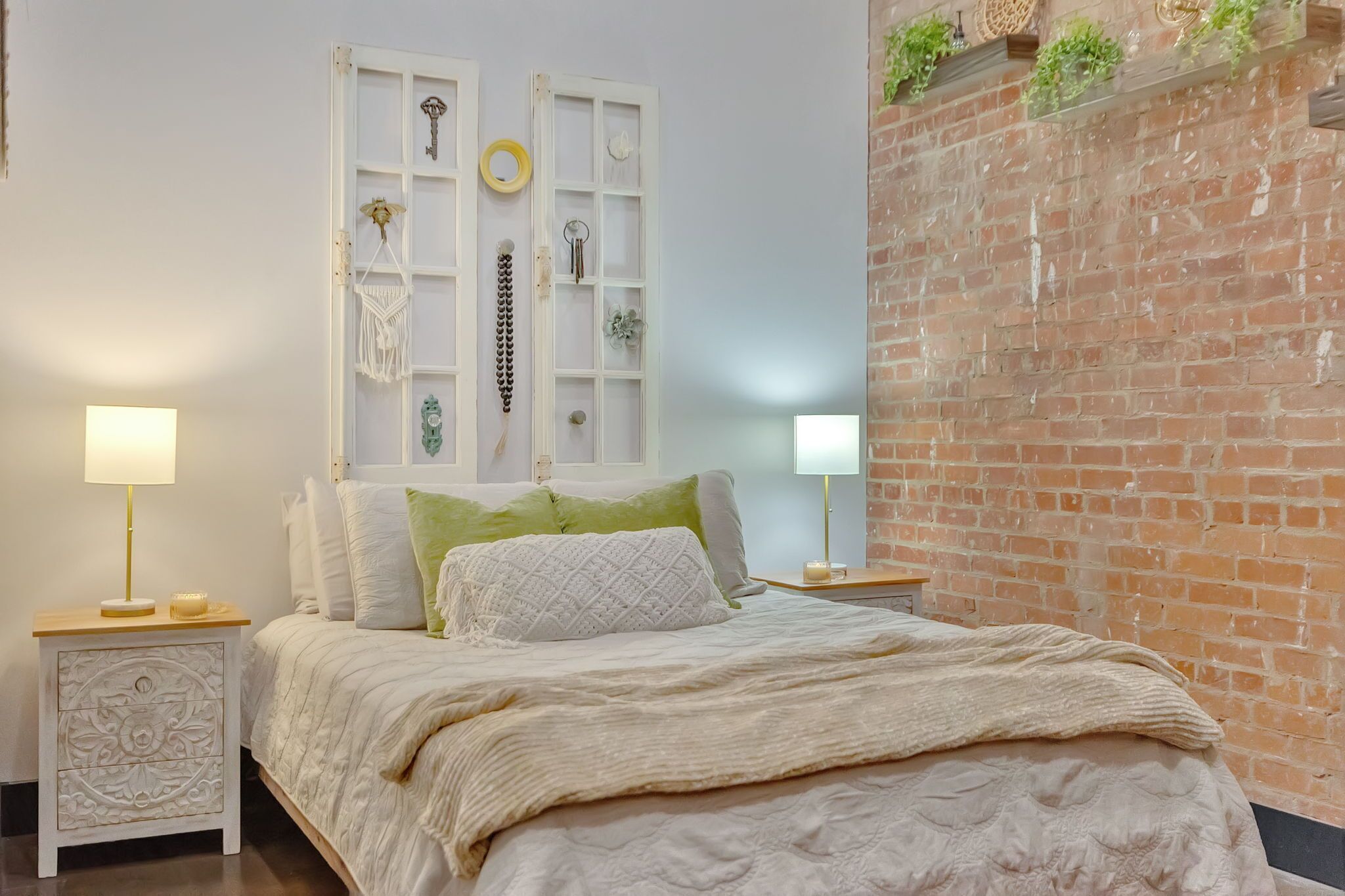 A bedroom with a bed, two bedside tables, a white wall, a brick wall, and a window with decorative items.