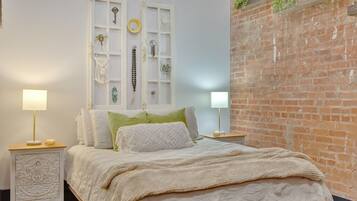 A bedroom with a bed, two bedside tables, a white wall, a brick wall, and a window with decorative items.