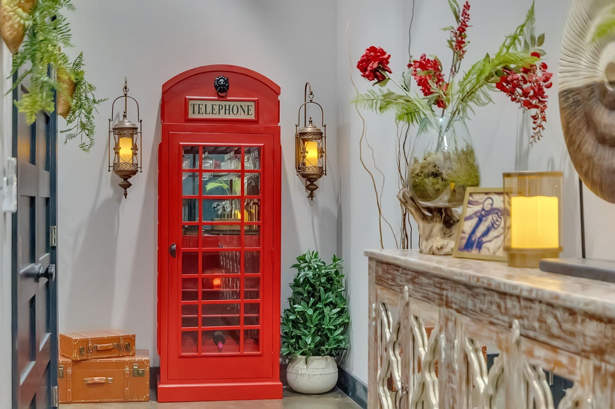 Une cabine téléphonique rouge avec une enseigne, deux lanternes fixées au mur, une plante en pot et un vase décoratif avec des fleurs.