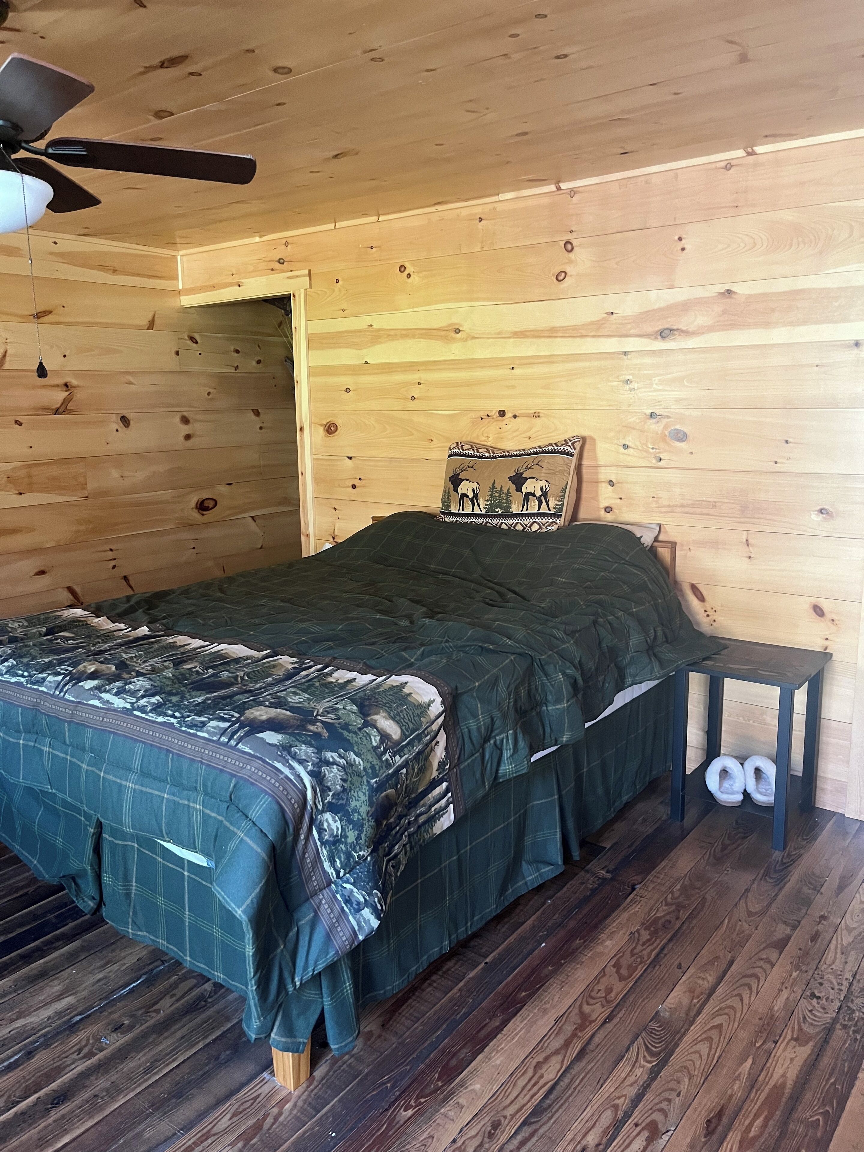 A cozy wooden room with a bed, a pillow with a deer design, a small table with slippers, and a ceiling fan.
