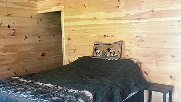 A cozy wooden room with a bed, a pillow with a deer design, a small table with slippers, and a ceiling fan.