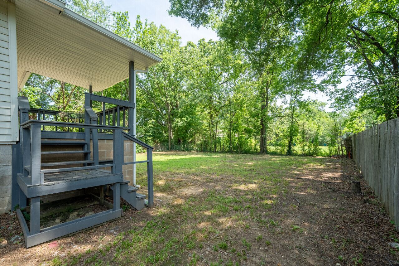 A covered porch with a wooden staircase leading to a deck, surrounded by a grassy yard and a wooden fence.