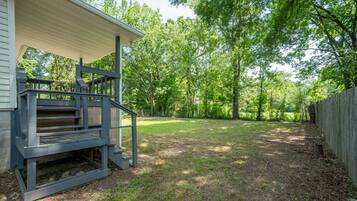 A covered porch with a wooden staircase leading to a deck, surrounded by a grassy yard and a wooden fence.