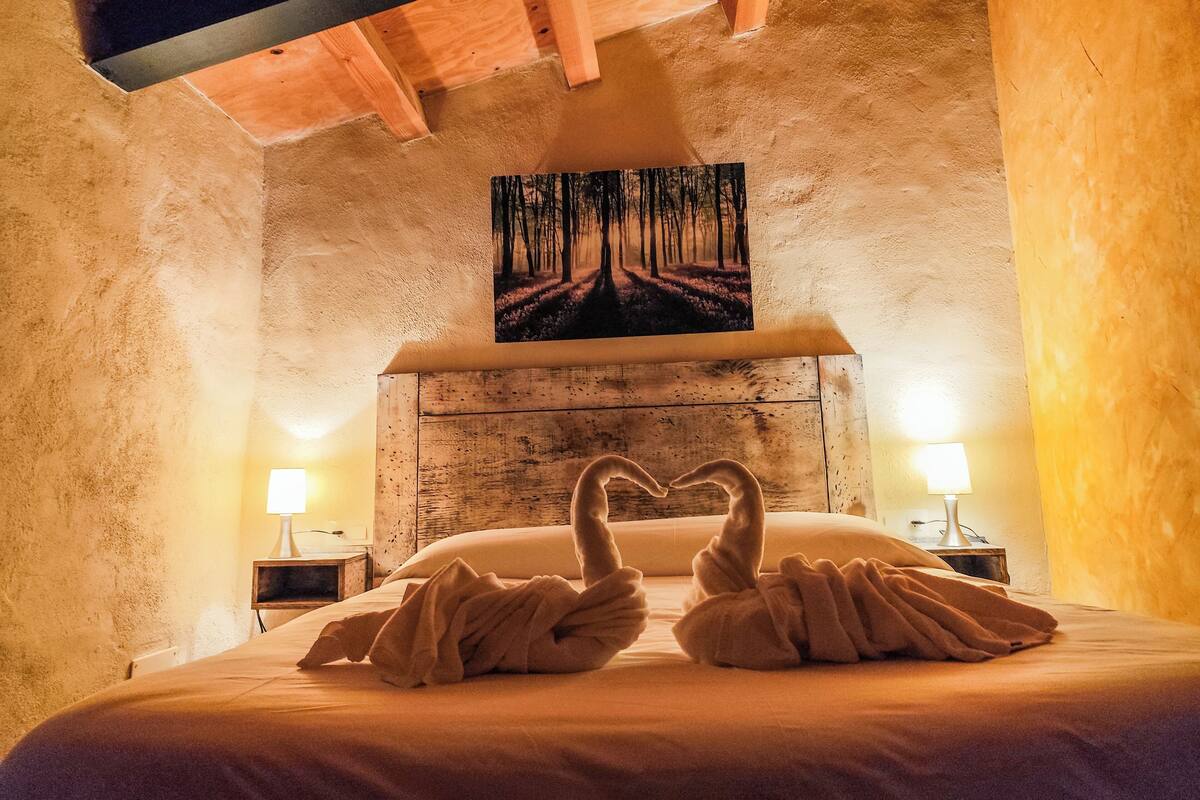 A cozy room with a bed, two nightstands, a painting of a forest, and a ceiling with exposed beams.