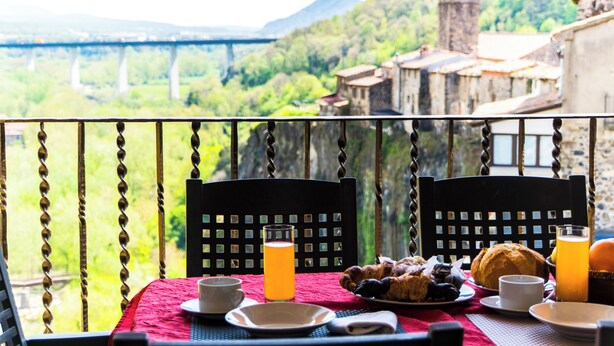Una mesa con alimentos para el desayuno, un vaso de jugo de naranja y una vista panorámica de un puente y follaje.