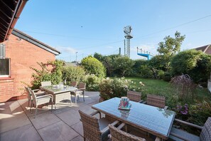A patio with wicker furniture and a glass table, surrounded by a garden with various plants and a brick wall. - Sea View 5* Property, Sleeps 10, Parking, Dogs (Whitby)