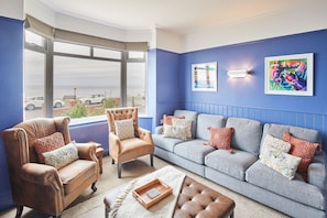 Una sala de estar con una pared azul, un ventanal grande, un sillón de cuero marrón, un sofá gris y una mesa de centro de madera.