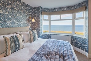 A bedroom with a large bed, a window with a view of the beach, and patterned wallpaper. - Sea View 5* Property, Sleeps 10, Parking, Dogs (Whitby)