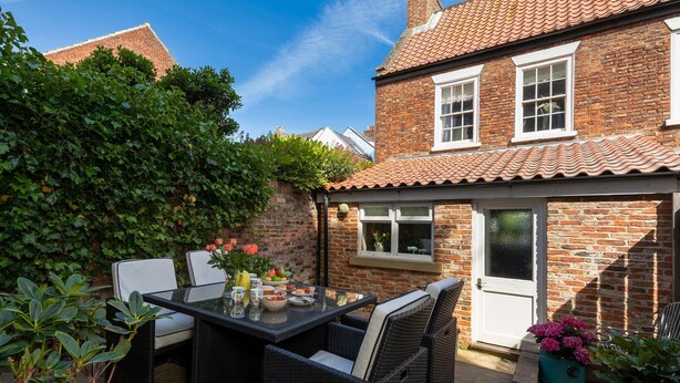 A brick house with a tiled roof, a patio with a table and chairs, and a window with a view of the sky.