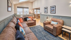 A spacious living area with a large sectional sofa, a smaller sofa, and a bench. There are framed pictures on the walls, a fireplace, and a kitchen area in the background.