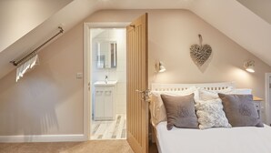 A bedroom with a sloped ceiling, a bed with multiple pillows, a bedside lamp, a heart-shaped wall decoration, and a bathroom with a mirror and sink.