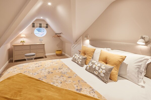 A bedroom with a bed, a dresser, a vanity, a round window, and a hanging light fixture.