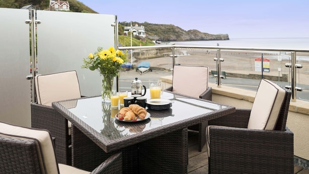 Une table à manger avec des aliments pour le petit-déjeuner, des chaises en osier et une vue sur la plage et les falaises.