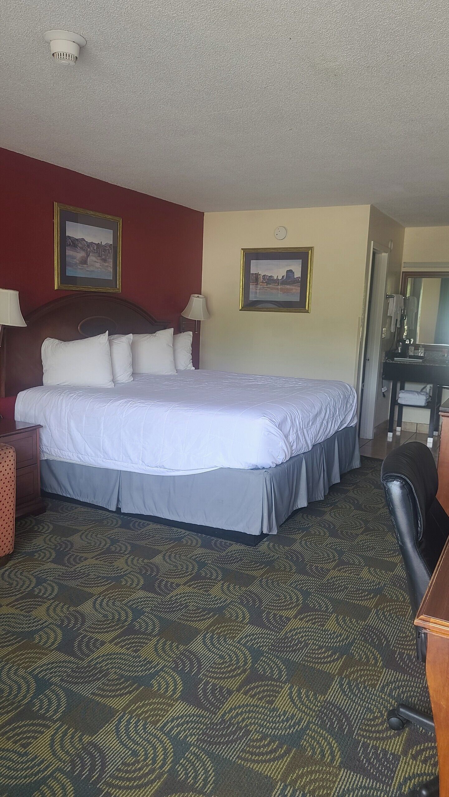 A hotel room with a large bed, a desk, a chair, and two framed pictures on the walls.